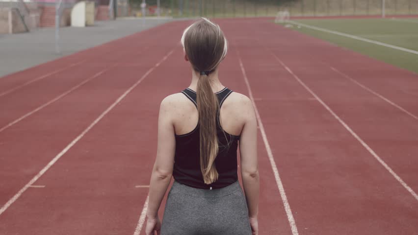 Female runner is standing on stadium track preparing to run, a view from back, slide up. Female athlete stands at start of stadium on street preparing for the race, the concept of aspiration and way