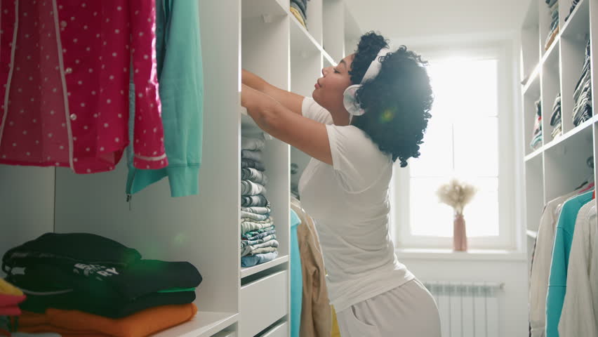 Portrait of African American woman with curly hair, having fun and dancing while listening music in headphones, organizing wardrobe. Maid female doing spring cleaning in walk in closet. Service staff Royalty-Free Stock Footage #1097995319