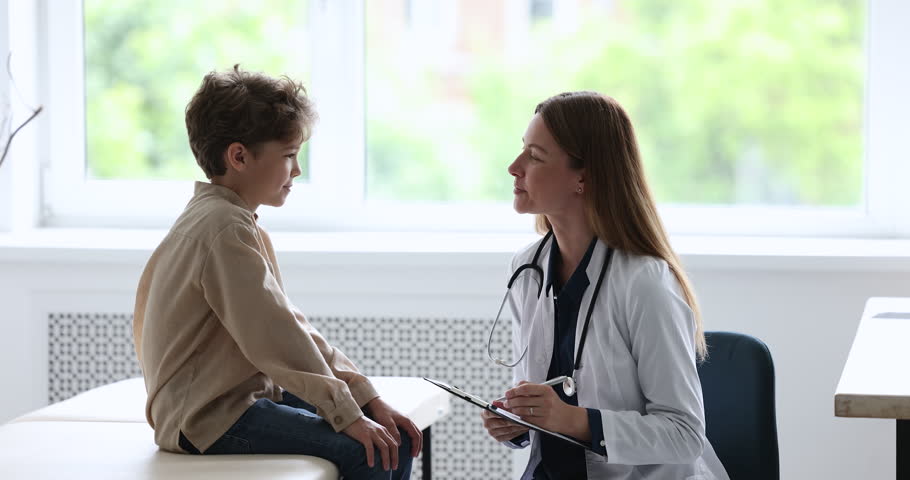 Young woman doctor pediatrician in white uniform ask illness symptoms, writes, filling form talks to little 8s boy patient during visit in clinic or hospital. Medicine, paediatric, health diagnostics Royalty-Free Stock Footage #1098002565