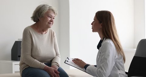 Female doctor in white coat listen to older woman tell about chronic disease, health complaints, affliction symptoms, therapist take notes, fill history form during patient visit. Nursing, elder care Video Stok