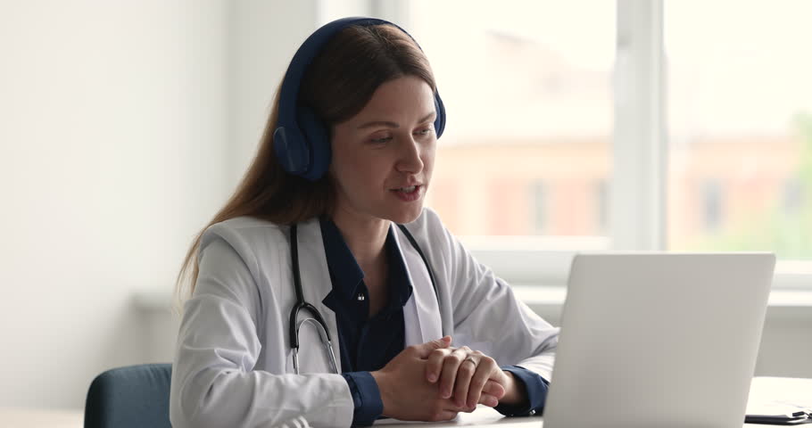 Young attractive woman general practitioner in white coat wear headphones sit at desk look at computer consulting to client distantly use videocall app. Virtual meeting event, wireless tech, medicare Royalty-Free Stock Footage #1098002597