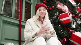 Young attractive blonde woman in red hat have video call conference meeting conversation on smartphone sitting near Christmas decoration on snowy city Happy smiling lady talking by smartphone outdoors