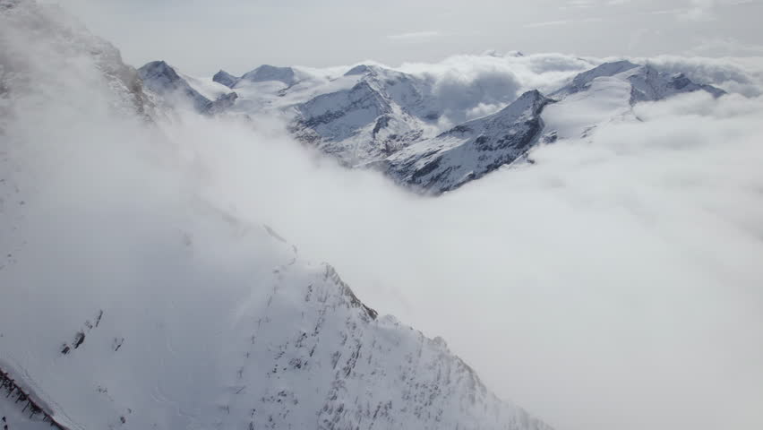 Mystic clouds surrounding icy and snow-covered mountains during sunny day in Austria - Aerial orbiting shot 