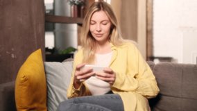 Soft dreamy portrait of charming blond woman gamer sitting on sofa relaxing alone and playing in online video games on smartphone holding in horizontal landscape mode at living room at home