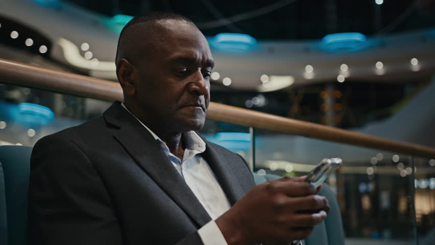 African businessman entrepreneur browsing cellphone smartphone indoors middle-aged man using mobile technology chatting free WI-FI connection indoors in company sitting at sofa browsing internet phone Royalty-Free Stock Footage #1098022595