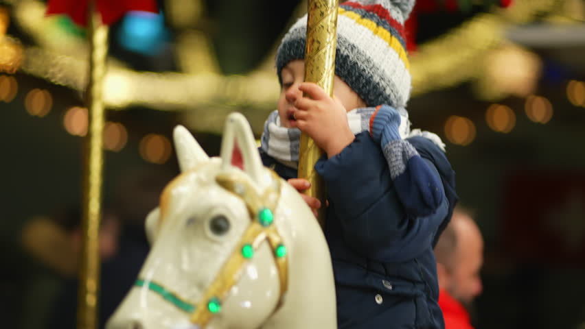 One happy little boy riding carousel in motion waving hello. Child in moving merry go round at amusement park wearing beanie and scarf during winter season. Christmas festivities