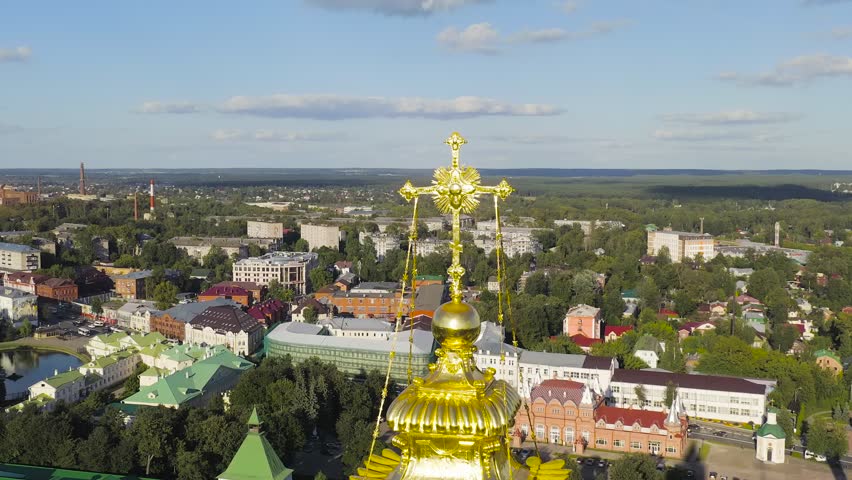 Sergiev Posad, Russia. The Trinity-Sergius Lavra is a fortified Orthodox monastery complex centered around a white stone bell tower crowned with a golden onion dome and cross. The camera pulls back from a close aerial view of the gilded dome to reveal the full monastery ensemble, including the tall five-tiered bell tower, the Trinity Cathedral with its distinctive golden and blue domes, and the surrounding white defensive walls with green rooftops. The complex sits amid dense summer greenery with the residential and industrial buildings of the town visible in the background under a bright sky with scattered clouds. The Lavra was inscribed as a UNESCO World Heritage Site in 1993 and its 88-meter bell tower remains one of the tallest in Russia. The warm late-afternoon light casts long shadows across the historic compound, emphasizing the golden surfaces and white stone architecture. The smooth aerial pullback captures the monastery