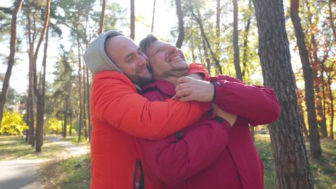 Happy LGBT male couple hugging looking away dating on sunny day in autumn park. Medium shot portrait of satisfied confident Caucasian gay men embracing standing on park alley smiling. Diversity Video Stok
