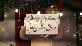 Happy holidays. Young smiling woman holding board with text Merry Christmas and Happy New Year, standing outdoors against building brick wall decorated with xmas led lights and firtree branches, video