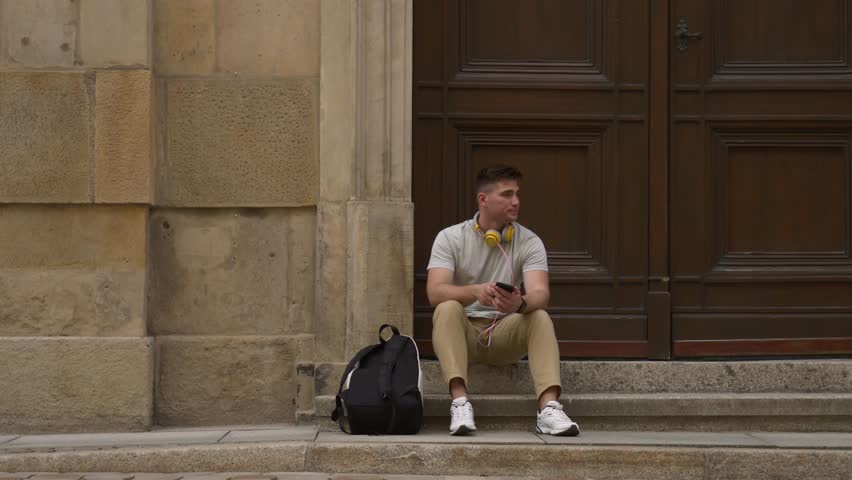 Student guy with mobile phone sits on stairs of University in Wroclaw, Poland