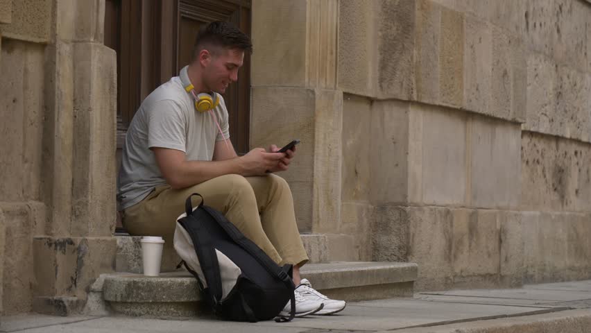Student guy with mobile phone sits on stairs of University in Wroclaw, Poland