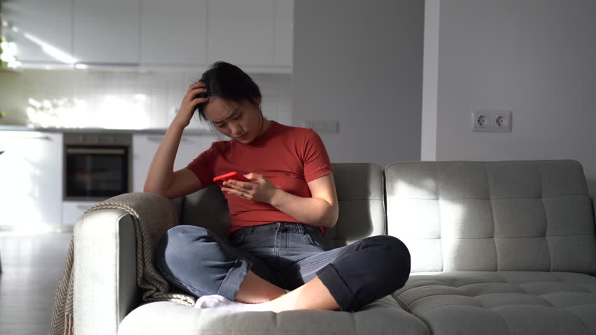 Sad emotional Asian girl throwing mobile phone away while breaking up with boyfriend via text. Upset anxious woman getting bad news on mobile phone, coping with break up, sitting on couch at home | Shutterstock HD Video #1098051215