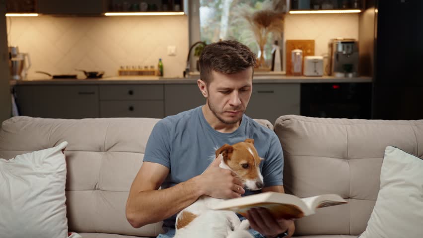 Man sitting on sofa in living room, reading paper book and stroking dog. Resting at home with pet, relaxation with book