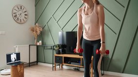 Woman is doing sports exercises in living room. Remote classes with fitness trainer at home, using laptop for video call. Woman in sports clothes does cardio on the floor