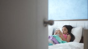 asian child addicted smartphone or kid girl sleep lying to pillow on bed and play watching mobile phone to hyperactive with parents secretly looking at daughter or baby behind ajar door in bedroom