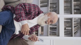 Smiling elderly man talks to his family on video call via smartphone resting in comfortable chair against rack with bookshelves. Bearded pensioner enjoys online communication at home 