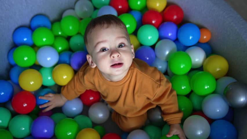 Adorable kid sitting in dry pool with balls looks up curiously into camera. Lovely boy wants to stand up but fails. Top view.