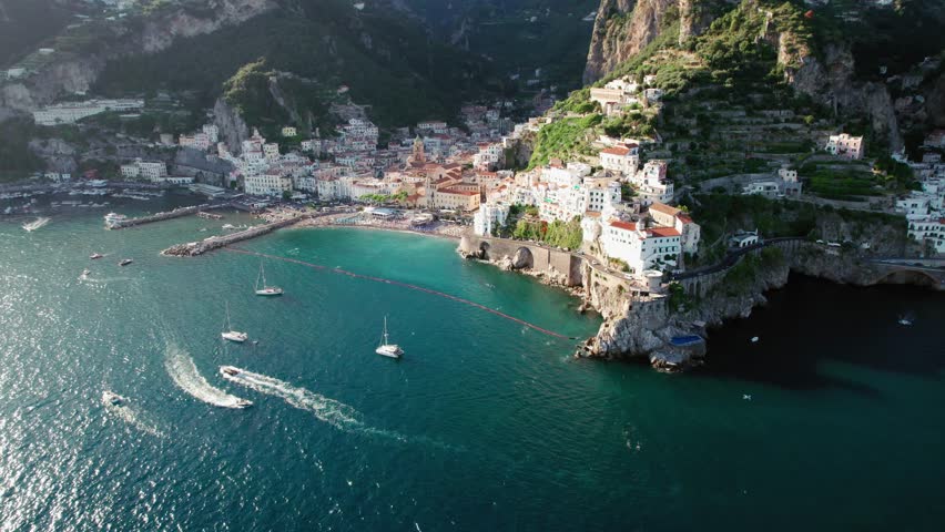 An extreme wide aerial panning around the busy port of Amalfi as boats sail in to the harbor and tourists lounge at beach Il Duoglio Spiaggia in Amalfi town along the famous Amalfi Coast in Italy. Royalty-Free Stock Footage #1098078385