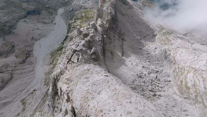 Breathtaking drone shot of rocky mountains with flying clouds and green valley in Dolomites - tilt up