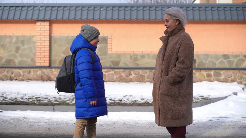 Side view positive mature mother and son gesturing high-five waving goodbye leaving. Wide shot Caucasian woman and schoolboy saying goodbye before school on winter morning walking away