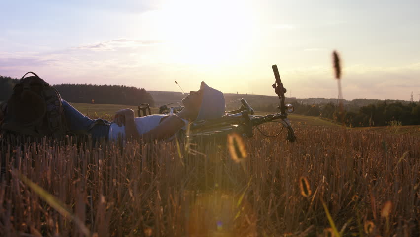 Funny teenager boy relax outdoors, lies near the bike at sunset in the field, teen boy getting ready for bed in the field, cinematic steadicam shot, rack focus
