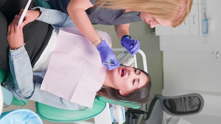 Vertical video Professional Woman Dentist Examines Female Patient with a Special Tool and Prescribes Treatment in a Modern Dental Clinic. Oral Hygiene. Concept of Healthcare and Medicine. Slow Motion Royalty-Free Stock Footage #1098084503