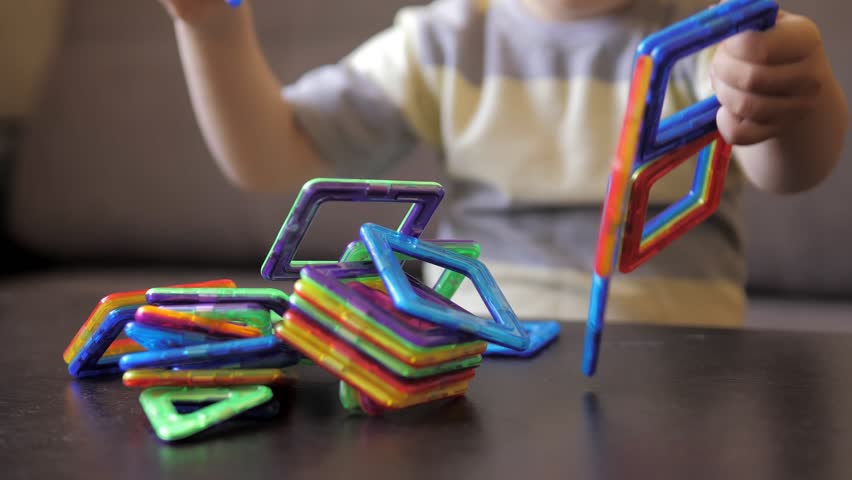 Cute 3 years old caucasian boy is sitting at the table on a sofa and playing a magnetic constructor building a house. Creativity development. Magnetic constructor. Carefree weekend