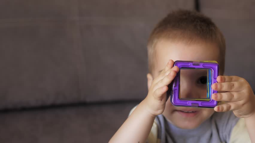 Cute 3 years old caucasian boy is sitting at the table on a sofa and playing a magnetic constructor building a house. Creativity development. Magnetic constructor. Carefree weekend