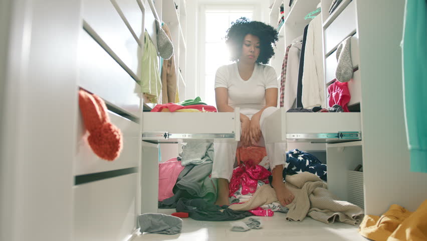 Cute African American girl trying to find needed dress by throwing out all clothes out of drawers in closet. Colorful clothes falling on floor creating mess in wardrobe. Nothing to wear concept 4K Royalty-Free Stock Footage #1098095359