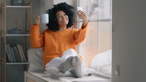 Happy woman drinking coffee in front of phone camera at window. Pretty girl making video on smartphone. African American woman posing for photo with cup of hot tea in living room interior slow motion