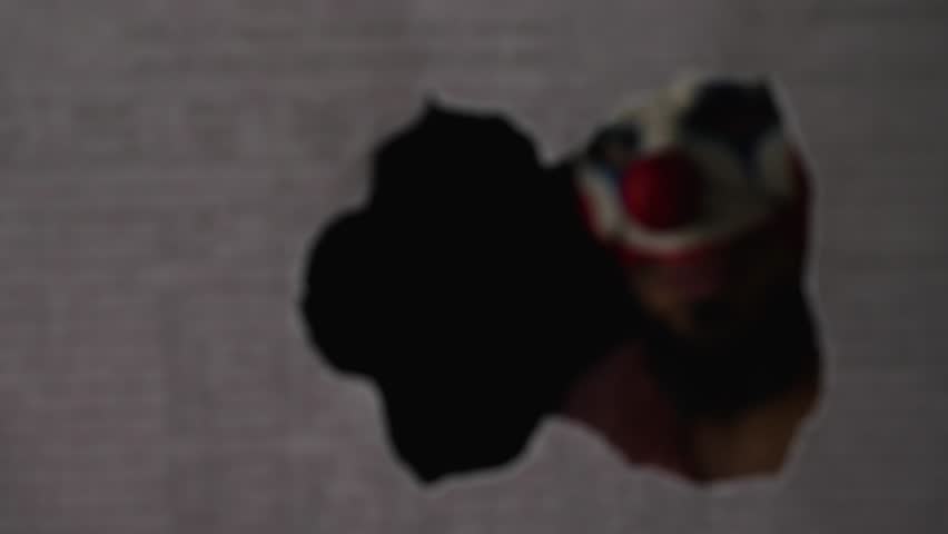 A scary clown looks into the camera through a hole in the wall. Close-up. The face of a man in a clown mask peering at his victim through a hole in the wall. Creepy clown maniac Royalty-Free Stock Footage #1098113893