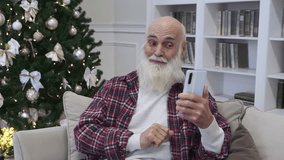 Elderly man talks to his family on video call via smartphone resting in comfortable chair against rack with bookshelves. Bearded pensioner enjoys online communication at home 