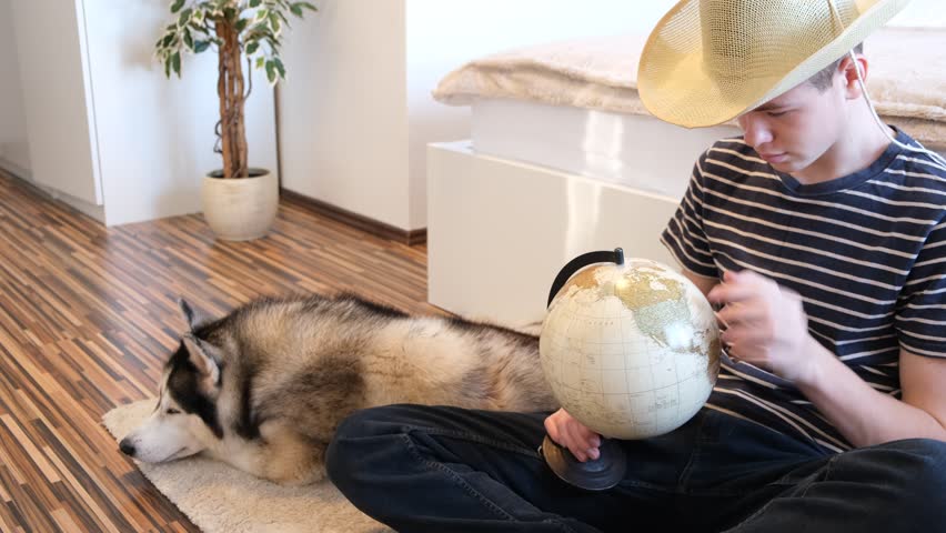 A teenage boy in a striped T-shirt, jeans and a hat sits in a room on the floor and turns a globe in his hand, a husky dog lies next to him. Top view closeup