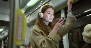 Side-view portrait of attractive happy caucasian female using public transport standing holding handrail watching videos on smartphone and wireless headphones. Charming young woman travelling by bus.