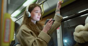 Side-view portrait of attractive happy caucasian female using public transport standing holding handrail watching videos on smartphone and wireless headphones. Charming young woman travelling by bus.