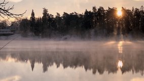 Smoke on the water. Sunrise. Beautiful winter lake in the foggy morning. Pines trees silhouetts forest in the distance. Sea bay or lake calm surface of the water and misty clouds float above it. 