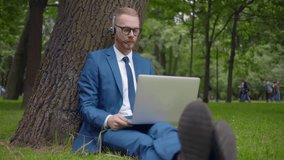 Male positive entrepreneur freelancer making online video call on laptop outdoors. Businessman in headphones have video conference on laptop computer in park. Realtime