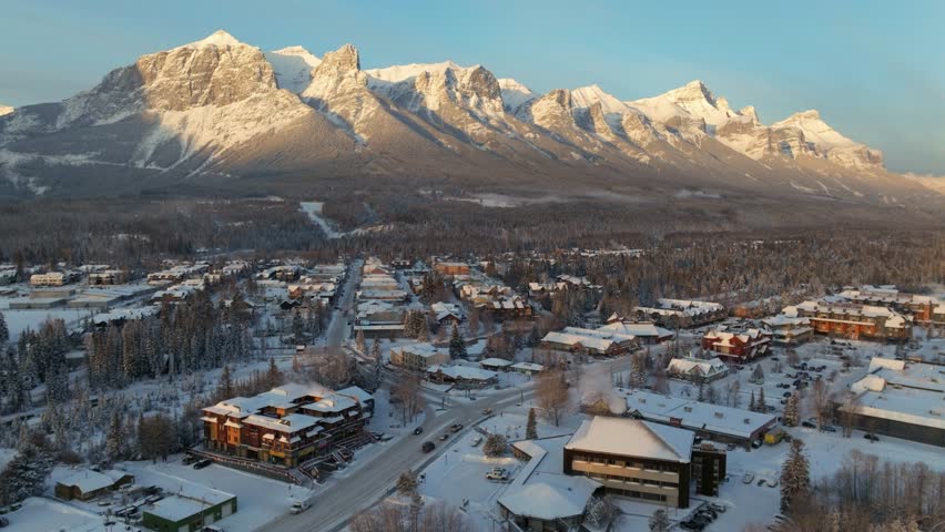 The amazing Canadian Rockies peer over the quaint town of Canmore Alberta. Golden hour sun hits the peaks. Aerial. Royalty-Free Stock Footage #1098138995