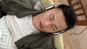 Vertical Video: view from above happy asian young man is singing into smarthpone while lying on the couch listening to pop songs from headphones in the living room at home.
