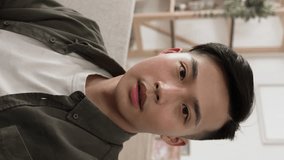 Vertical Video: cheerful asian guy looking and waving hands at the camera is talking with finger pointing while making a video call in a modern cozy home interior.