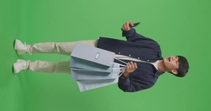 Full View Of An Asian Shopping Man With Shopping Bags Having Video Call On Mobile Phone While Standing In Front Of Green Screen
