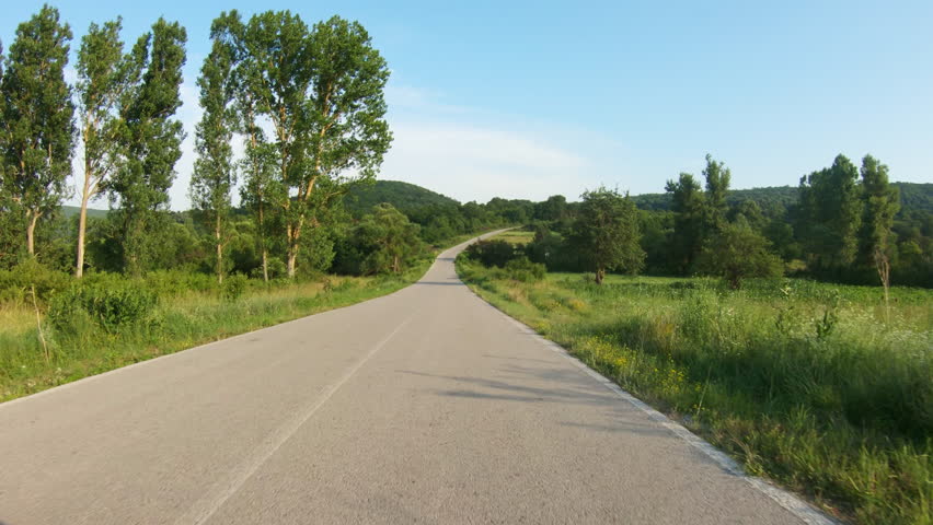 A slow car or motorcycle ride uphill on a country road surrounded by meadows and trees. Blue sky, summer time. POV shot