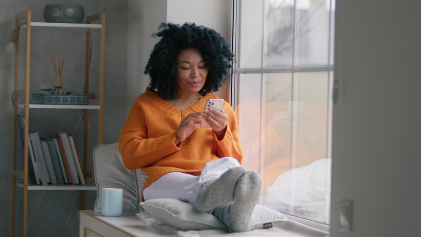 Happy African American model using smartphone device at home. Smiling woman with curly black hair blogger subscribing new social media on cell phone, buying internet, ordering products online in apps Royalty-Free Stock Footage #1098163611