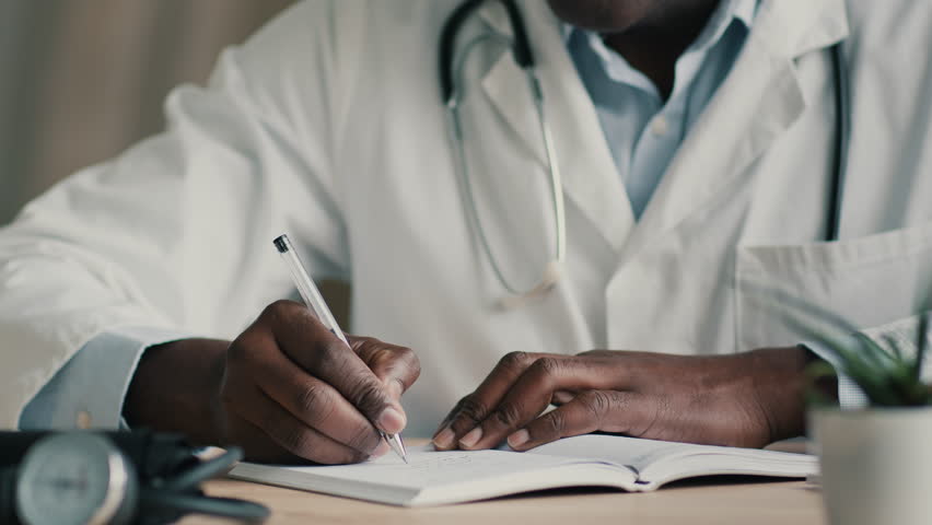 Close up view african man doctor general practitioner medic doing paperwork writing recipe at desk filling checklist health symptoms or med insurance write medical notes in notebook planning workday | Shutterstock HD Video #1098168259