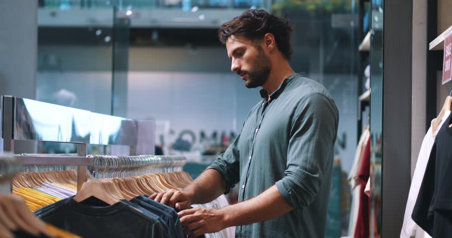 Attractive bearded multi-ethnic macho man in dark-green casual shirt, shopping for t-shirts in fashionable men's clothing store, inspecting differents shirts, price tags and the quality of fabric Royalty-Free Stock Footage #1098173371