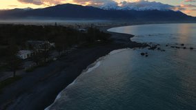 Aerial drone shot of sea with lapping waves and snow capped mountain background houses orange dusk sky new Zealand