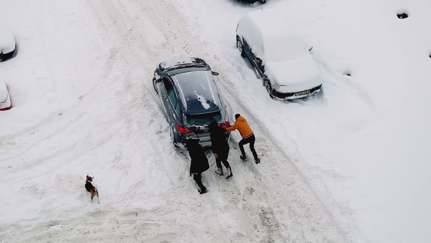 People are pushing a car out of snowdrifts in the courtyard of a house on the road. Ice under the snow on the road, winter tires. Windstorm