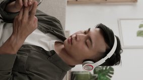 Vertical Video: happy asian man with headphones is moving his body to the rhythm on the couch after choosing and playing songs on the smartphone at a bright home interior.