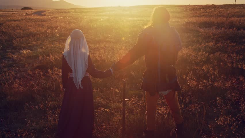 A man and a woman in medieval costumes twitches against the backdrop of a sunset in the mountains. View from the back. Between them is a sword that is stuck in the ground