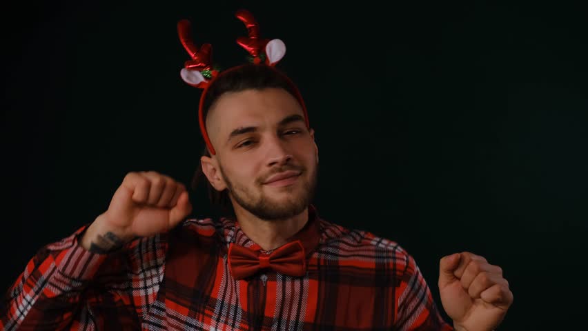 Young Caucasian bearded man with red Christmas deer horns wears plaid shirt and bow tie, portrait on dark studio background. Man dances and enjoys Christmas and New Year holidays.